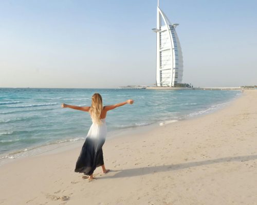 A carefree woman in a flowing dress strolls along the Dubai beach with Burj Al Arab in view.